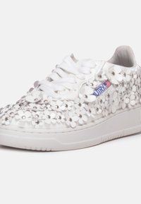 Zapatilla blanca con aplique floral y acentos metálicos, suela acolchada, punta redonda y diseño de cordones tradicional.