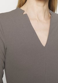 Šedé šaty s výstřihem do V a zářezem, vyrobené z hladké látky. Mají dlouhé rukávy a jemný šev uprostřed.