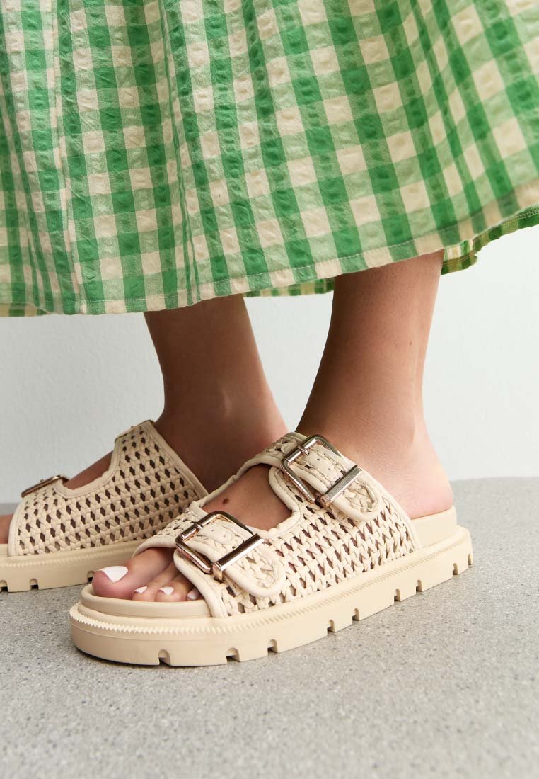Sandalias deslizantes de tejido beige con dos hebillas metálicas, suela gruesa y material de goma texturizada, contra un fondo a cuadros verde y blanco.