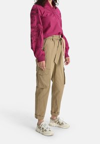 Camicia fucsia con bottoni e ricami floreali sulle maniche, abbinata a pantaloni cargo color khaki e sneakers beige con accenti grigi.