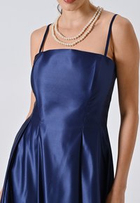 Marineblauwe satijnen jurk met een aansluitend lijfje, dunne bandjes en een geplooide rok. Een opvallende parelketting voegt contrast toe aan de halslijn.