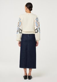 Elfenbeinfarbener Pullover mit gestickten Motiven an den Ärmeln, darunter Blätter und Sterne. Kombiniert mit einem dunkelblauen Jeans-Midirock. Schwarze Loafers mit Hardware-Details.