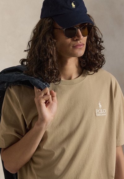 Νέος άνδρας με σγουρά μαλλιά φοράει σκούρα γυαλιά ηλίου και μπλε σκούφο, μπεζ μπλουζάκι Polo Ralph Lauren, κρατώντας τζιν μπουφάν στον ώμο.