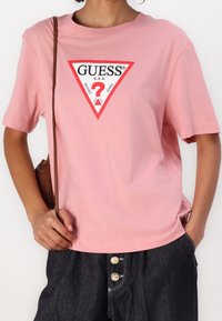 Persona vistiendo una camiseta rosa con el logo de Guess en rojo y blanco, pantalones de mezclilla oscuros con botones dorados y una correa de bolsa marrón.