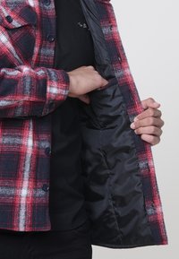 Punane ja tumesinine ruuduline flanellist särgitagi siniste nuppudega ja musta nailonvoodriga; näitab sisetaskut ja kontrastset materjali.