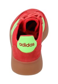 Sneaker rossa con tomaia in suede, accenti verde neon e suola in gomma marrone. Presenta lacci e un logo embossato sul tallone.