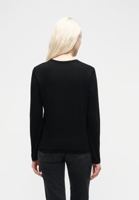 Γυναίκα με μακριά ξανθά μαλλιά που φοράει απλό μαύρο πουλόβερ με μακριά μανίκια και σκούρο γκρι τζιν, όρθια μπροστά σε λευκό φόντο, γυρισμένη από την πλάτη.