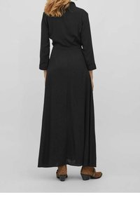 Černé dlouhé šaty s přiléhavým střihem, dlouhými rukávy a hladkým povrchem, doplněné hnědými botami na podpatku. Jednoduchý, elegantní design.