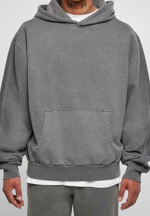 Person trägt einen locker sitzenden grauen Kapuzenpullover mit einer Fronttasche und passende graue Hosen, darunter ist der Saum eines weißen Hemdes sichtbar.