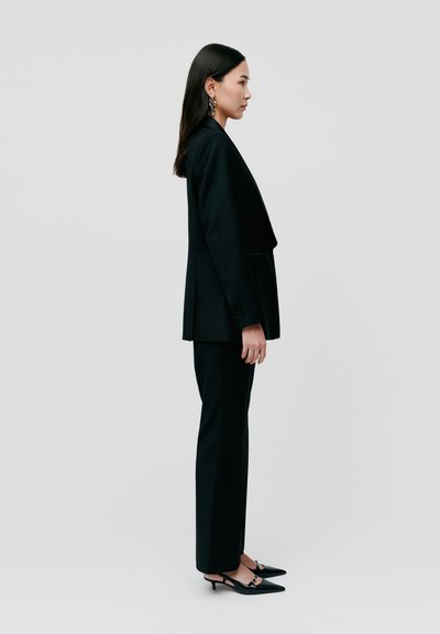 Black tailored suit with long blazer and slim-fit trousers, paired with pointed black heels. Smooth fabric and structured silhouette.