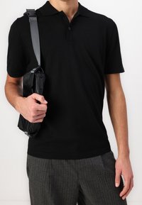 Man in een zwart polo shirt met korte mouwen en grijze gestreepte broek, die een kleine zwarte schoudertas met een brede schouderband vasthoudt.