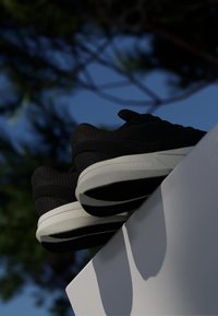 Crne sportske cipele s bijelim potplatima na bijeloj površini, na nejasnoj vanjskoj pozadini s drvećem i plavim nebom.