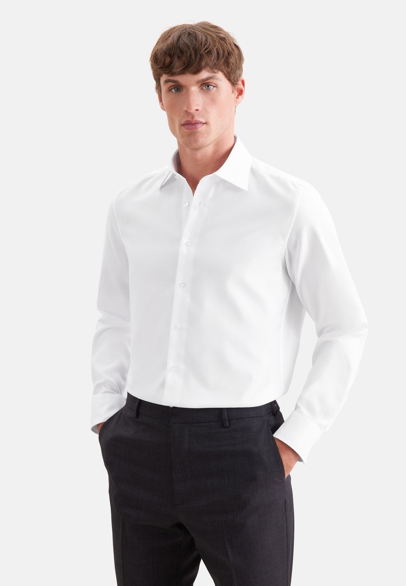 Homme portant une chemise blanche cintrée et un pantalon noir, debout les mains dans les poches devant un fond clair uni.