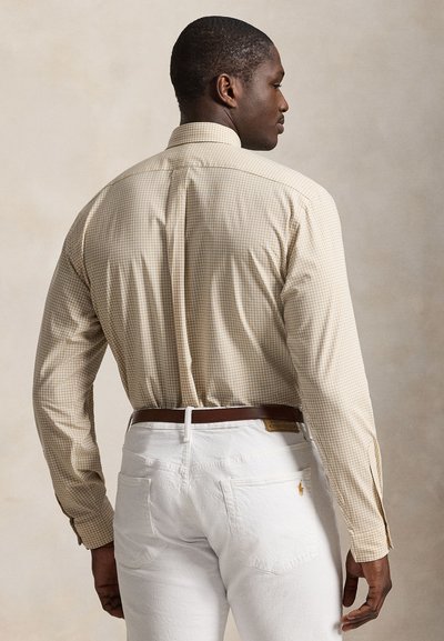 Homme de dos portant une chemise beige à carreaux à manches longues rentrée dans un pantalon blanc avec une ceinture marron sur un fond uni.