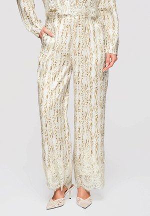Kvinde iført creme- og beige mønstrede vidde bukser med blondekant kombineret med matchende blondefarvet top og cremefarvede mesh-spidse hæle.