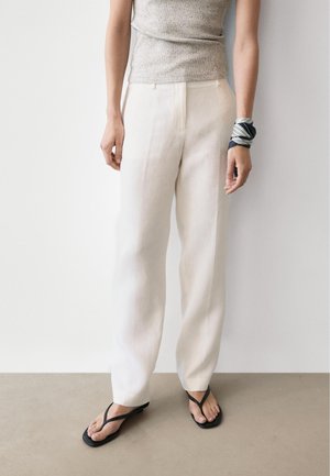 Kvinde iført hvide bukser med høj talje, grå ærmeløs top, sorte klipklappere og sort-hvidt stribet håndledssjal, stående op ad hvid væg.