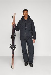 Crna topla ski jakna s teksturiranim dizajnom, džepovima na patent zatvarač i kapuljačom. Tamne hlače i skije s vezovima također su vidljive.