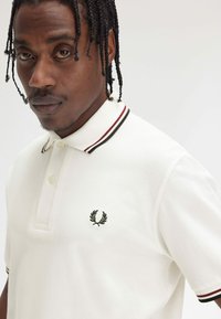 Polo bianco con tessuto testurizzato, colletto dettagliato con strisce rosse e verdi, e un emblema ricamato sul petto sinistro.