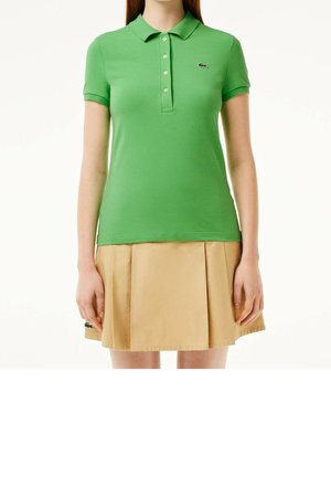 Kvinde iført en grøn poloshirt med korte ærmer med et lille logo og et beige plisseret skørt, der står mod en hvid baggrund.