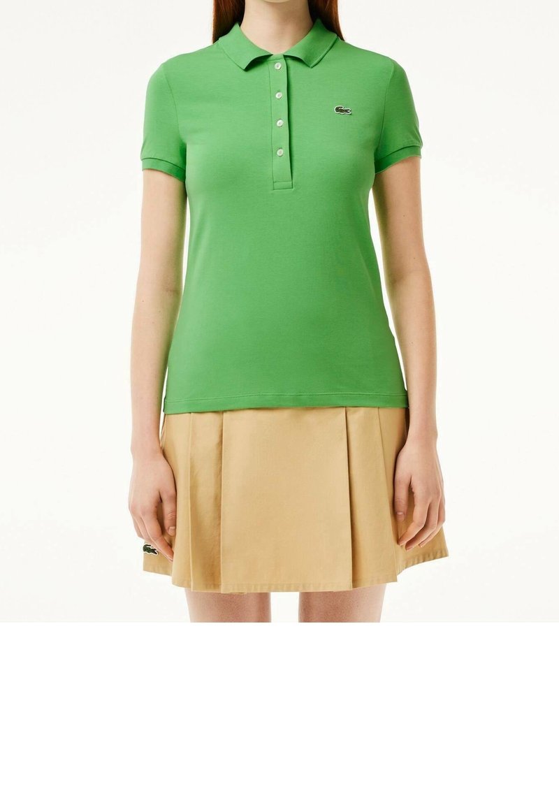 Femme portant un polo vert à manches courtes avec un petit logo et une jupe plissée beige, debout devant un fond blanc.