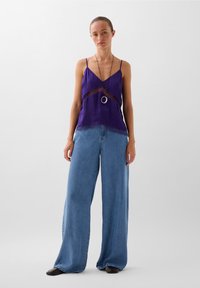 Camiseta de satén púrpura con detalles de encaje en el dobladillo, combinada con unos vaqueros de pierna ancha en azul claro. Modelo lleva un collar largo tipo colgante.