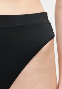 Κοντινό πλάνο ενός ατόμου που φοράει μαύρο μαγιό με ψηλή μέση, δείχνοντας τη μέση και το πάνω μέρος του μηρού σε απλό φόντο.