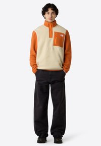 Fleece pullover in crème en oranje, met een ritskraag, borstzak met rits, een relaxte fit en zwarte cargobroek.