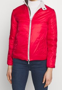 Červená prošívaná bunda s lesklou texturou, zipovým zapínáním a světlým límečkem na patentky. Má dlouhé rukávy a přiléhavý střih.