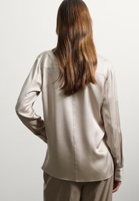 Beige satin Langarmhemd mit lockerer Passform, Knopfmanschetten und glatter Textur. Von hinten gezeigt mit einfachen Schneiderdetails.