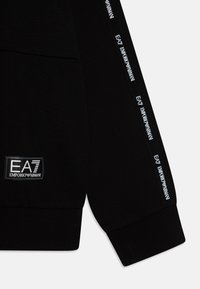 Černá mikina s texturovanou tkaninou, žebrovými manžetami a logem s nápisem "EA7 EMPORIO ARMANI". Doplňky zahrnují značkovou pásku podél rukávů.