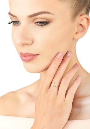 Gold ring with a T-shaped design and gemstone accent, worn on a hand resting near a face with smooth skin and neutral nails.