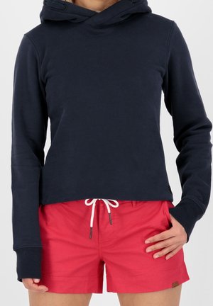 Osoba ubrane w granatową bluzę z kapturem i jaskrawoczerwone szorty ze białym sznurkiem, stojąca z jedną ręką w kieszeni, a drugą opartą na udzie.