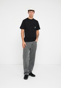 Črn bombažni T-majica s prsno žepom, v kombinaciji s sivimi jeans hlačami. Model nosi črne čevlje in belo kapo. Preprost, sproščen videz.