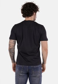 Camiseta negra de manga corta en algodón suave con cuello redondo, combinada con jeans azules. Presenta un sutil logotipo en la manga izquierda.