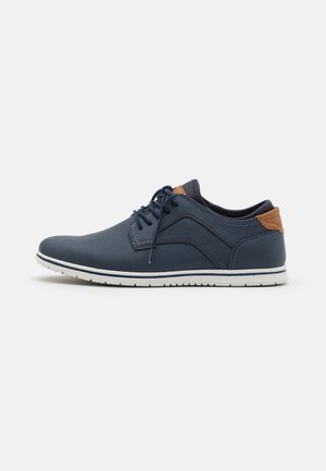 Men's lace-up shoes in navy blue synthetic material with a contrasting brown heel accent, textured sides, and white rubber sole.