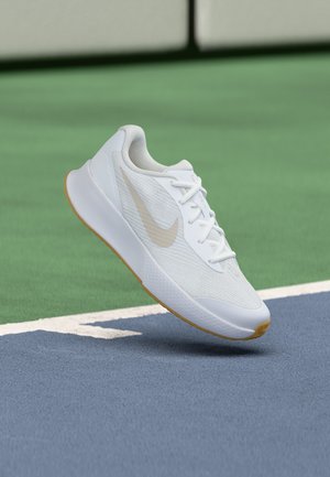 Biela teniska Nike s béžovým logom a gumovou podrážkou vznášajúca sa nad modrozeleným povrchom tenisového kurtu blízko bielej čiary.