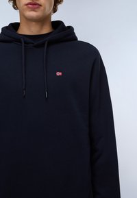 Mørkeblå hoodie i blødt stof med en frontkangaroo-lomme, der har et lille broderet rødt og hvidt flag. Justerbar snoretræk til hætten.