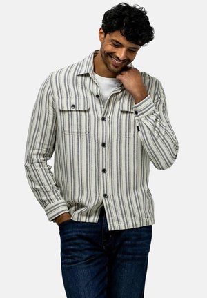 Man glimlacht met zijn hoofd naar beneden gekanteld, draagt een wit-zwart verticaal gestreept overhemd over een wit T-shirt en donkerblauwe jeans.