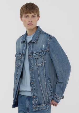 Giacca di denim blu chiaro con una vestibilità rilassata, due tasche sul petto, cuciture a vista e dettagli invecchiati. Indossata sopra un maglione blu chiaro.