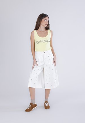 Kvinde iført gul tanktop med teksten "CaféNoir", hvide vide cropped bukser med små mønstre, beige sokker og brune sneakers, stående.