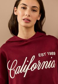 Bordeaux hættetrøje med "California" i hvid kursiv og "EST 1989" i en simpel skrifttype, lavet af blødt stof.