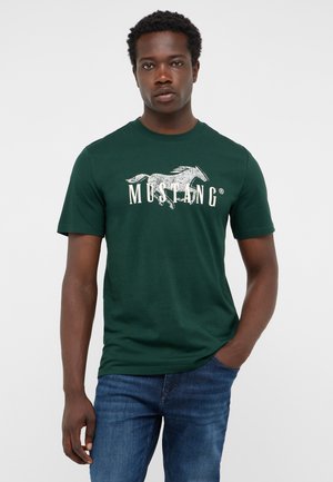 Young man with dark skin and short curly hair wearing a green Mustang logo t-shirt and blue jeans standing with one hand in pocket.