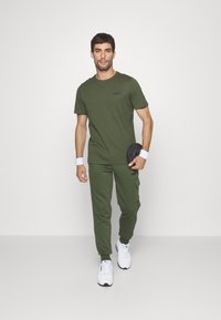 Conjunto deportivo en verde oliva que incluye una camiseta de manga corta y pantalones estilo cargo, combinados con zapatillas deportivas blancas y muñequeras de color gris oscuro.