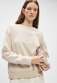 Beżowa bluza z fakturowanym, okrągłym haftowanym wzorem, z prążkowanymi mankietami i luźnym krojem. Modelka trzyma dopasowaną, jasnobeżową torbę.
