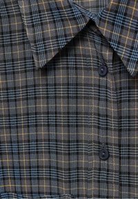 Gros plan sur un col de chemise à carreaux foncés boutonné, avec des lignes bleues et jaunes formant un motif à carreaux.