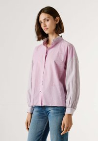 Roza in bela srajca z navpičnimi črtami, z razpadajočim ovratnikom in balon rokavi, narejena iz lahkega blaga. Ima zapenjanje na gumbe in subtilno blagovno znamko.