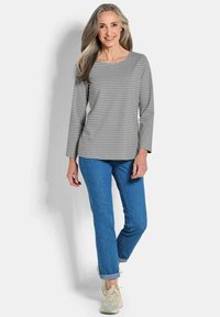 Glunderende vrouw met lang grijs haar, gekleed in een grijs gestreept shirt met lange mouwen, blauwe jeans en beige sneakers, staand tegen een witte achtergrond.