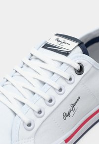 Baskets blanches Pepe Jeans London avec lacets blancs, logo noir sur le côté et la languette, et bande rouge et bleue sur la semelle.