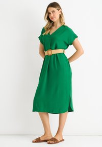 Abito verde con scollo a V e maniche corte, stretto in vita da una larga cintura intrecciata color sabbia. Il tessuto ha una superficie liscia e un design fino al ginocchio.