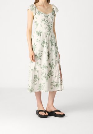Žena v bílé midi šatech se zeleným květinovým vzorem, krátkými rukávy, předním rozparkem a černých platformových žabkách stojící na bílém pozadí.